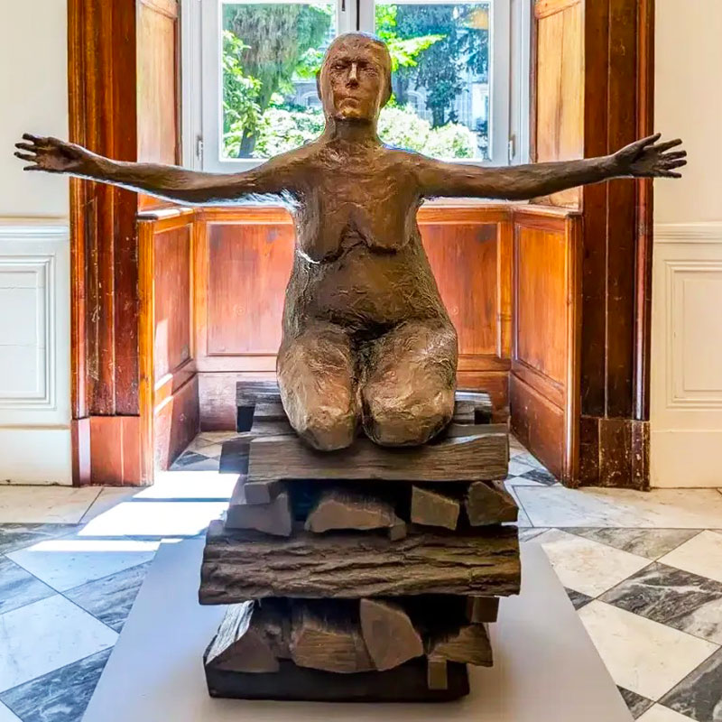 Elle représente une figure féminine nue, agenouillée, les bras grands ouverts dans une posture qui évoque à la fois l'offrande, la soumission et la résilience face au sacrifice. Elle repose sur un socle imitant des bûches de bois empilées. Fidèle à la démarche artistique de Kiki Smith, cette œuvre explore la vulnérabilité du corps humain, la condition féminine ainsi que les notions de martyre, d'histoire et de spiritualité (faisant souvent écho à des figures historiques ou mythologiques sacrifiées comme Jeanne d'Arc ou les sorcières menées au bûcher). Cette pièce majeure fait partie d'une édition très limitée (trois exemplaires originaux) et a notamment été présentée en France lors de rétrospectives majeures consacrées à l'artiste, comme à la Monnaie de Paris ou actuellement au MO.CO. de Montpellier.