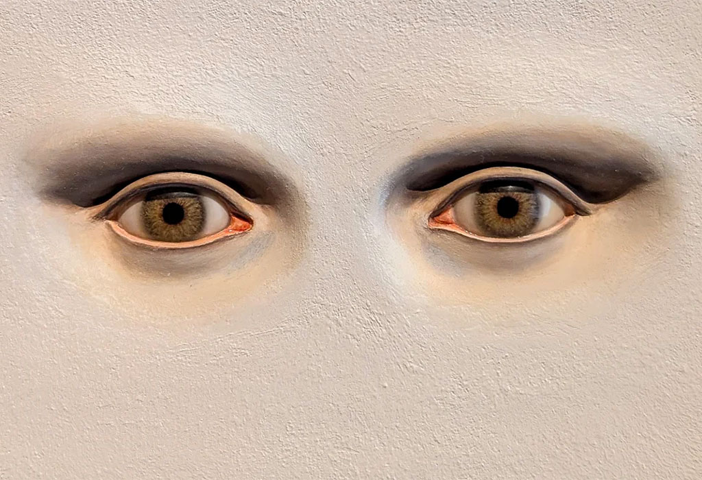 Cette image montre un détail saisissant d'une sculpture contemporaine en bas-relief de l'artiste française Serena Carone, représentant une paire d'yeux hyperréalistes encastrés directement dans la texture d'un mur blanc. « Ce que je vois », plâtre, peinture, 2010. H. 10 ; L. 15 cm