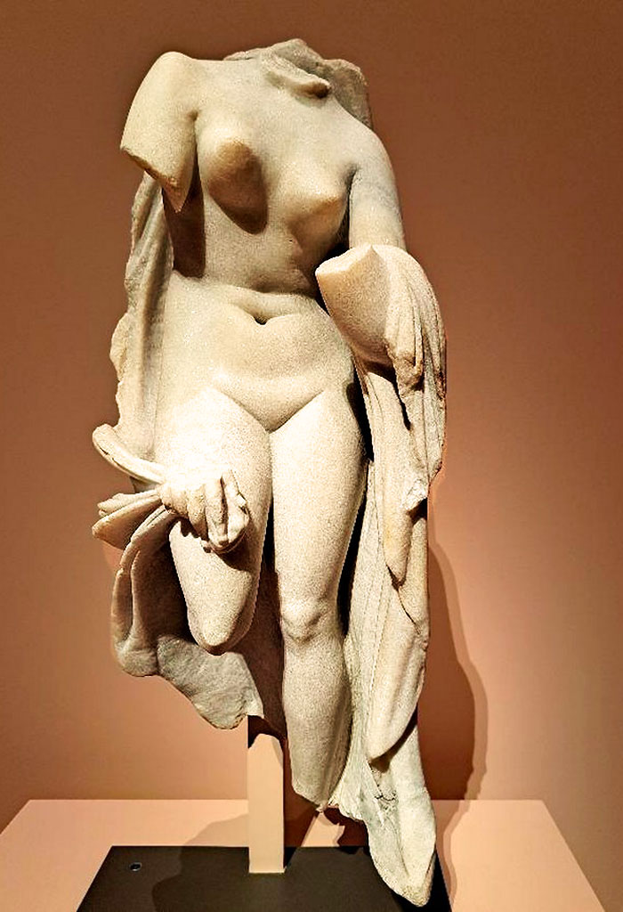Statuette antique d'Aphrodite (Vénus), la déesse de l'amour, de la beauté et de la séduction. Il s'agit d'une représentation classique d'Aphrodite sortant du bain, souvent inspirée des modèles hellénistiques dérivés de l'Aphrodite de Cnide de Praxitèle. Elle est représentée partiellement dénudée, retenant de ses mains un drapé (himation) tombant le long de son corps.
