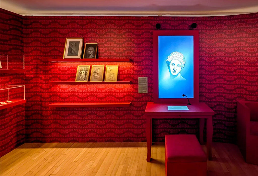 Cette image illustre un dispositif de médiation culturelle innovant basé sur l'intelligence artificielle au musée départemental Arles antique. Développée en collaboration avec l'entreprise spécialisée Ask Mona, cette installation immersive présente un avatar numérique animé de la célèbre Vénus d'Arles.